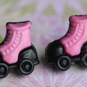 May include: A pair of pink and black roller skate earrings. The skates are made of plastic and have a black base with pink wheels.