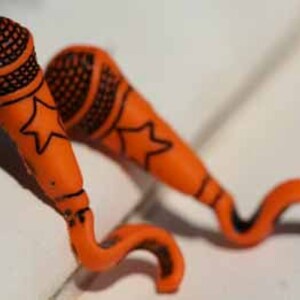 May include: A pair of orange microphone-shaped earrings with black star and microphone designs. The earrings are made of polymer clay and have a curved shape.