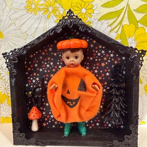May include: A doll wearing an orange pumpkin costume with a matching pumpkin hat, standing inside a black decorative house-shaped frame. The background features a black star pattern, a small black cat, a red mushroom, and a black tree.
