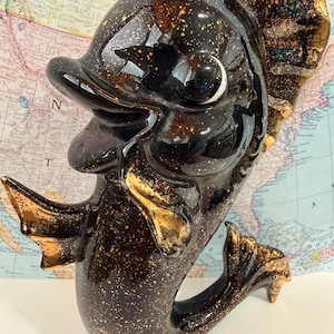 May include: A black ceramic fish figurine with gold speckles and accents. The fish has a smiling expression, large eyes, and detailed fins. The figurine is likely a decorative item for a home.