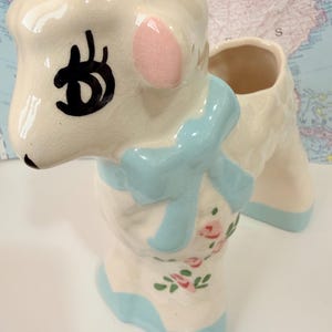 May include: A cream-colored ceramic lamb planter featuring a glossy finish with a crackle texture. The lamb has black painted eyes, pink ears, a light blue bow around its neck, and small pink floral accents with green leaves on its body.
