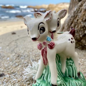 May include: A ceramic figurine of two white deer with brown spots, standing on a green base with brown and red accents. The deer have large eyes and are facing forward.