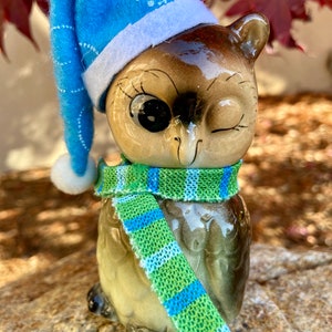 May include: A ceramic owl figurine with a blue winter hat and a green and blue scarf. The owl has one eye closed and is winking. The hat has a white trim and a snowflake design. The scarf is wrapped around the owl's neck.