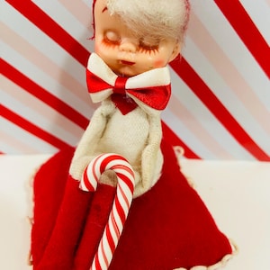 May include: A vintage Christmas doll with closed eyes, wearing a red bow tie and a red headband with a candy accent. The doll is holding a candy cane and sitting on a red cushion. The background has red and white stripes.