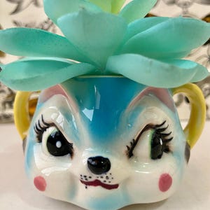 May include: A whimsical ceramic planter shaped like a winking bunny face, painted in blue, white, and pink. The planter has yellow handles and holds a faux succulent with light blue-green leaves. A fun and unique decorative item.