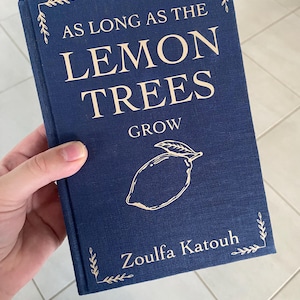May include: A navy blue book titled "AS LONG AS THE LEMON TREES GROW" with a lemon illustration. The author's name, Zoulfa Katouh, is printed below the illustration. Decorative leaf accents are in the corners.