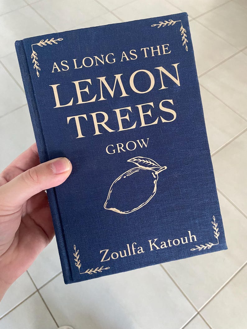 May include: A navy blue book titled "AS LONG AS THE LEMON TREES GROW" with a lemon illustration. The author's name, Zoulfa Katouh, is printed below the illustration. Decorative leaf accents are in the corners.