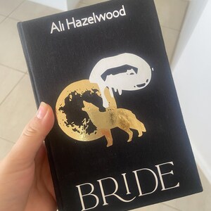 May include: A black book titled "BRIDE" by Ali Hazelwood. The cover features a gold illustration of a wolf and moon, with a white abstract design. The author's name is in white text at the top.