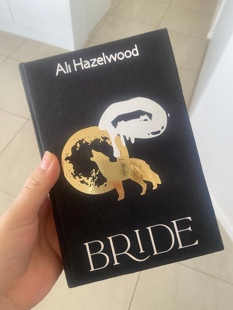May include: A black book titled "BRIDE" by Ali Hazelwood. The cover features a gold illustration of a wolf and moon, with a white abstract design. The author's name is in white text at the top.