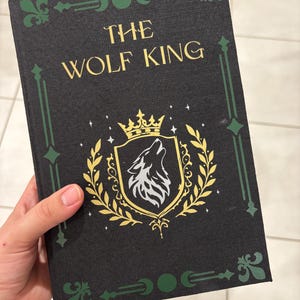 May include: A hardcover book titled "The Wolf King" with a black cover. The cover features gold lettering, a gold and white wolf crest, and green decorative accents. The book is held by a hand.