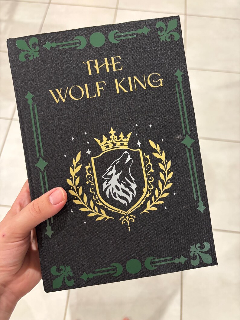 May include: A hardcover book titled "The Wolf King" with a black cover. The cover features gold lettering, a gold and white wolf crest, and green decorative accents. The book is held by a hand.