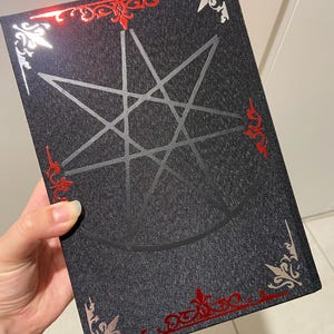 May include: A dark gray book with a silver pentagram design on the cover. The book has red and silver decorative accents around the edges. The book is held by a hand.