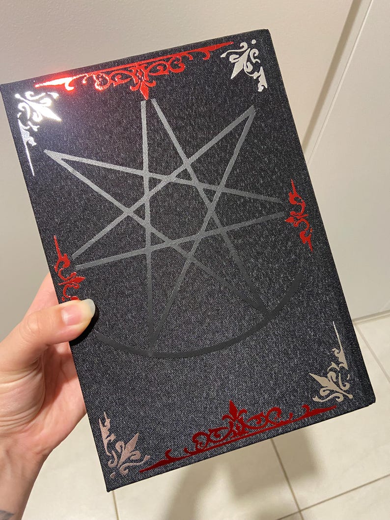 May include: A dark gray book with a silver pentagram design on the cover. The book has red and silver decorative accents around the edges. The book is held by a hand.