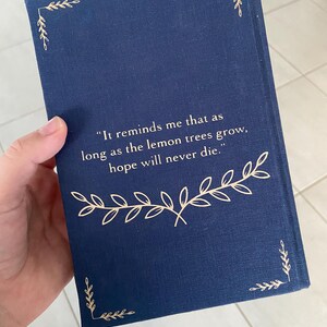 May include: A navy blue book cover with gold leaf accents. The cover features the quote, "It reminds me that as long as the lemon trees grow, hope will never die." Decorative gold leaf corner designs and a gold leaf floral design are also present.
