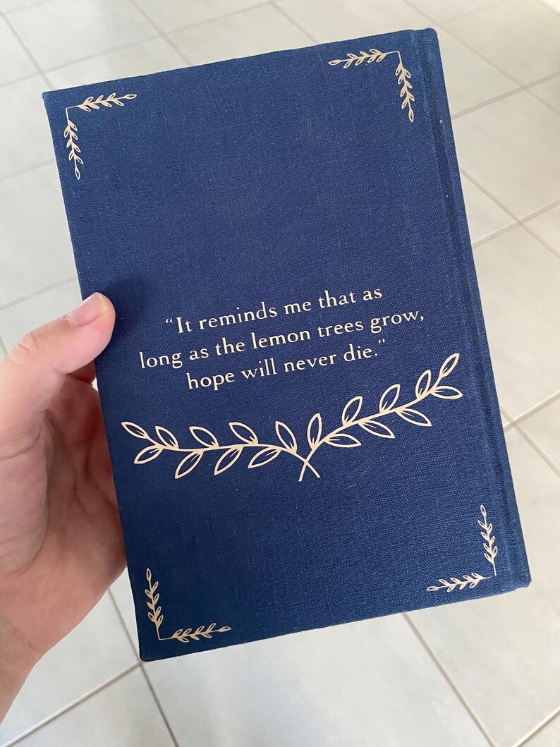 May include: A navy blue book cover with gold leaf accents. The cover features the quote, "It reminds me that as long as the lemon trees grow, hope will never die." Decorative gold leaf corner designs and a gold leaf floral design are also present.