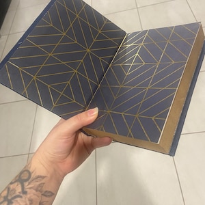 May include: Open navy blue book with gold geometric pattern on the inside covers. The book's edges are gold. The book is open, revealing the patterned interior. The cover is a solid navy blue.