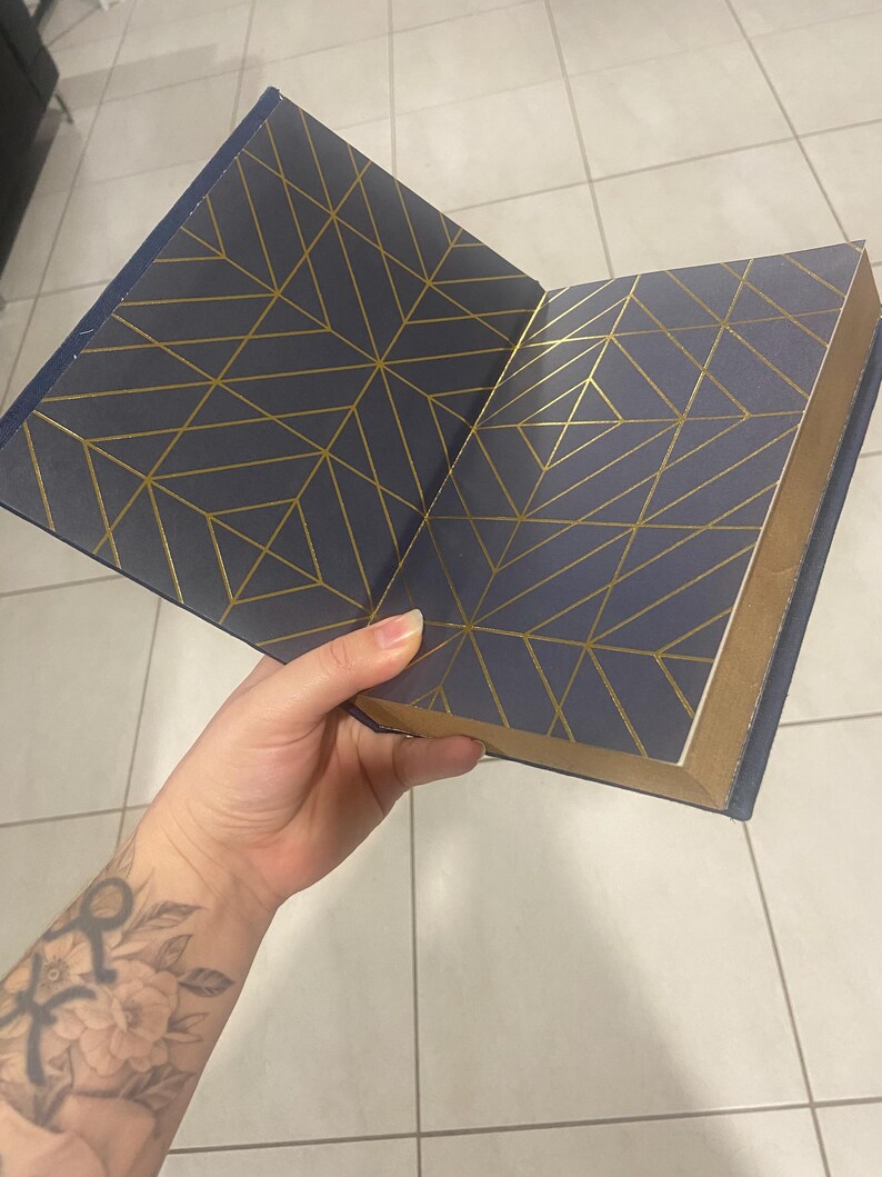 May include: Open navy blue book with gold geometric pattern on the inside covers. The book's edges are gold. The book is open, revealing the patterned interior. The cover is a solid navy blue.