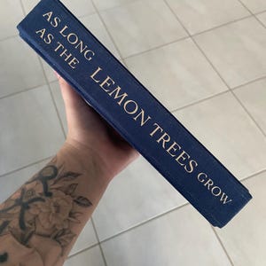 May include: A navy blue book with gold lettering that reads "AS LONG AS THE LEMON TREES GROW". The book is held at an angle, showcasing the title on the spine. The background is a light-colored tiled surface.