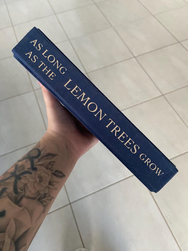 May include: A navy blue book with gold lettering that reads "AS LONG AS THE LEMON TREES GROW". The book is held at an angle, showcasing the title on the spine. The background is a light-colored tiled surface.