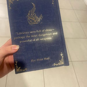 May include: A navy blue book with gold accents and text. The cover features a gold flame design and the quote: "Libraries were full of ideas&mdash;perhaps the most dangerous and powerful of all weapons." The book is inscribed "For Miss May."