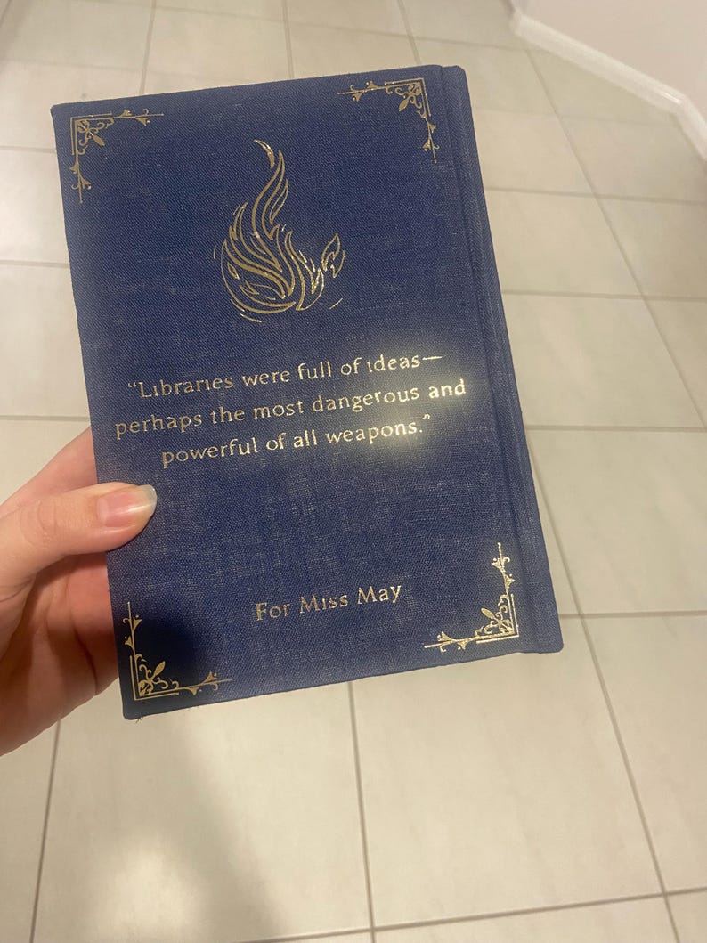May include: A navy blue book with gold accents and text. The cover features a gold flame design and the quote: "Libraries were full of ideas&mdash;perhaps the most dangerous and powerful of all weapons." The book is inscribed "For Miss May."
