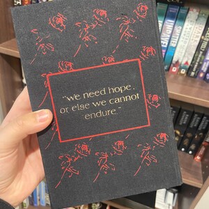May include: A black book cover with red rose illustrations and the quote, "we need hope, or else we cannot endure." The book is held in front of a bookshelf filled with other books.