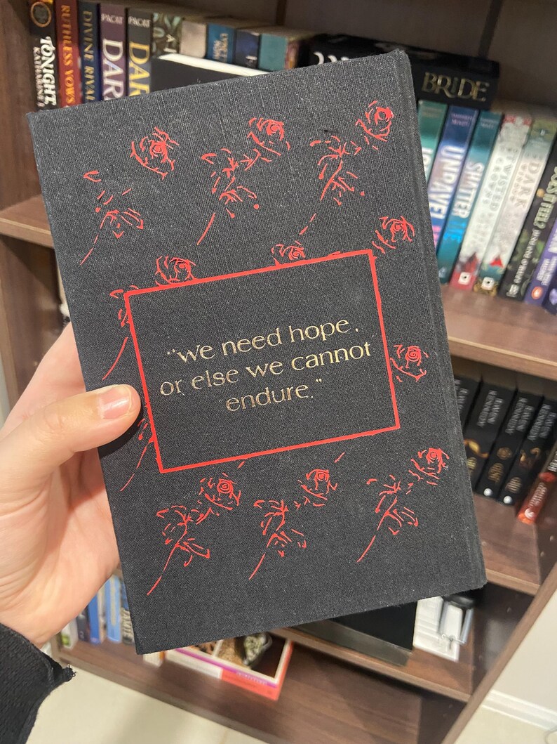 May include: A black book cover with red rose illustrations and the quote, "we need hope, or else we cannot endure." The book is held in front of a bookshelf filled with other books.