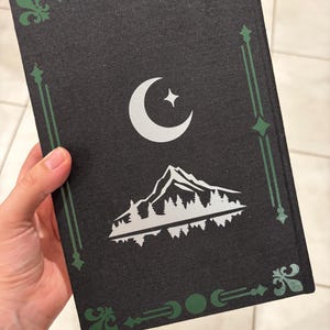 May include: Black book cover with a silver crescent moon and star design, and a mountain and forest scene. The cover has green decorative borders and accents. The book is held in a hand.