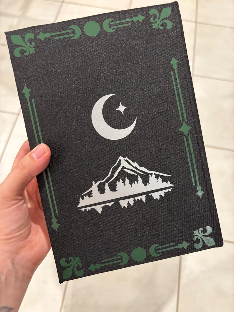May include: Black book cover with a silver crescent moon and star design, and a mountain and forest scene. The cover has green decorative borders and accents. The book is held in a hand.