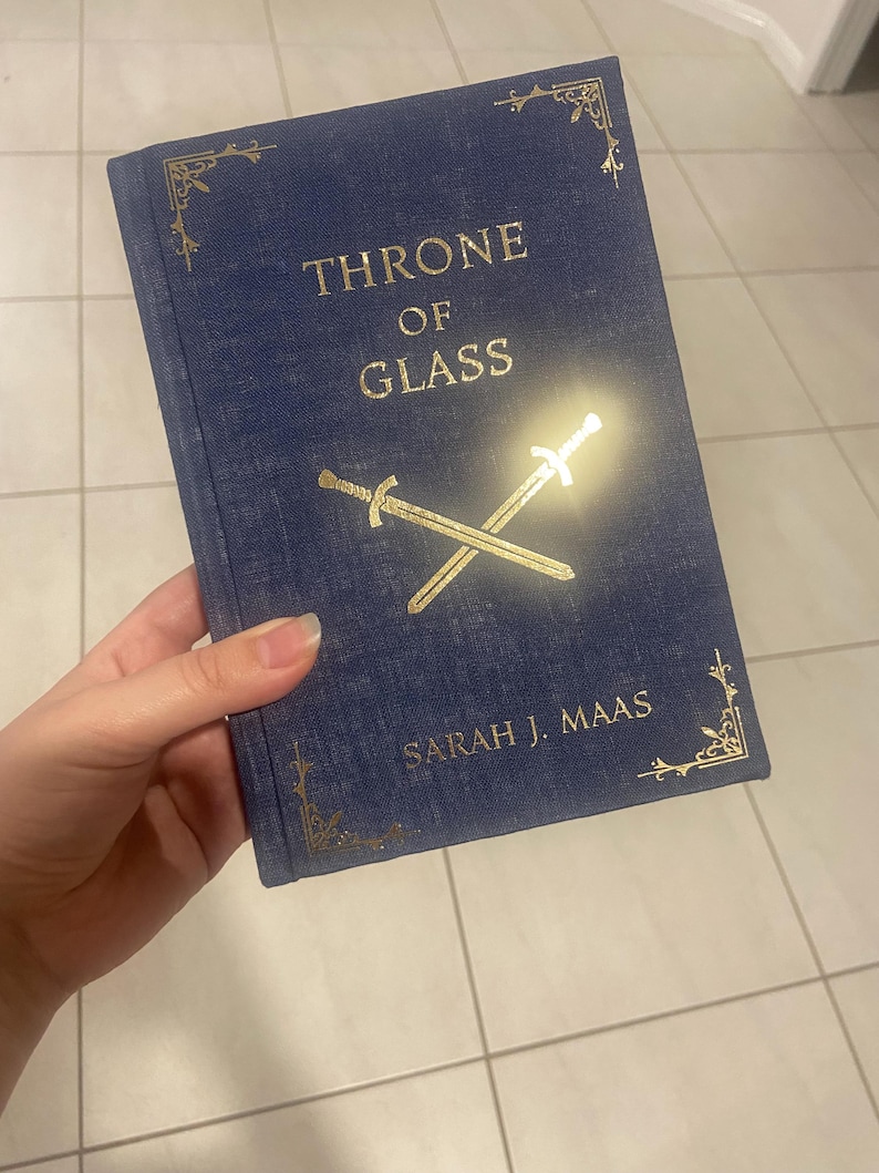 May include: A hardcover book titled "THRONE OF GLASS" by Sarah J. Maas. The book has a blue cover with gold lettering and crossed swords. Decorative gold corner designs add detail to the book's cover.