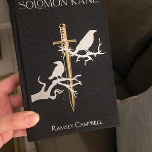 May include: A black book titled "SOLOMON KANE" by Ramsey Campbell. The cover features a gold sword and two white birds perched on branches. The book is held in a person's hand.
