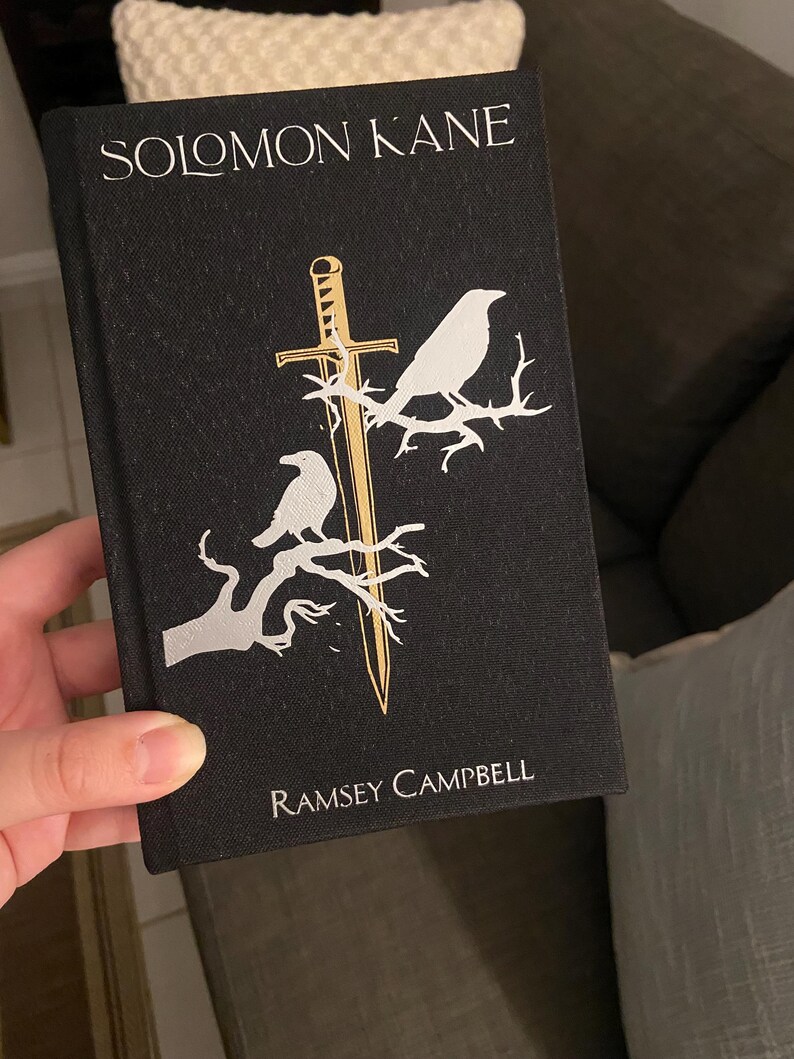 May include: A black book titled "SOLOMON KANE" by Ramsey Campbell. The cover features a gold sword and two white birds perched on branches. The book is held in a person's hand.