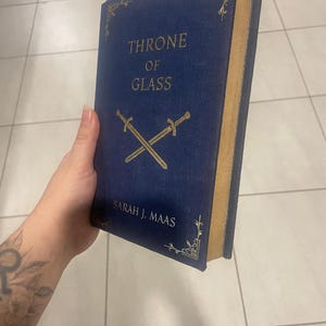 May include: A hardcover book titled "THRONE OF GLASS" by Sarah J. Maas. The book has a dark blue cover with gold lettering and decorative corner designs. Two crossed swords are featured below the title.