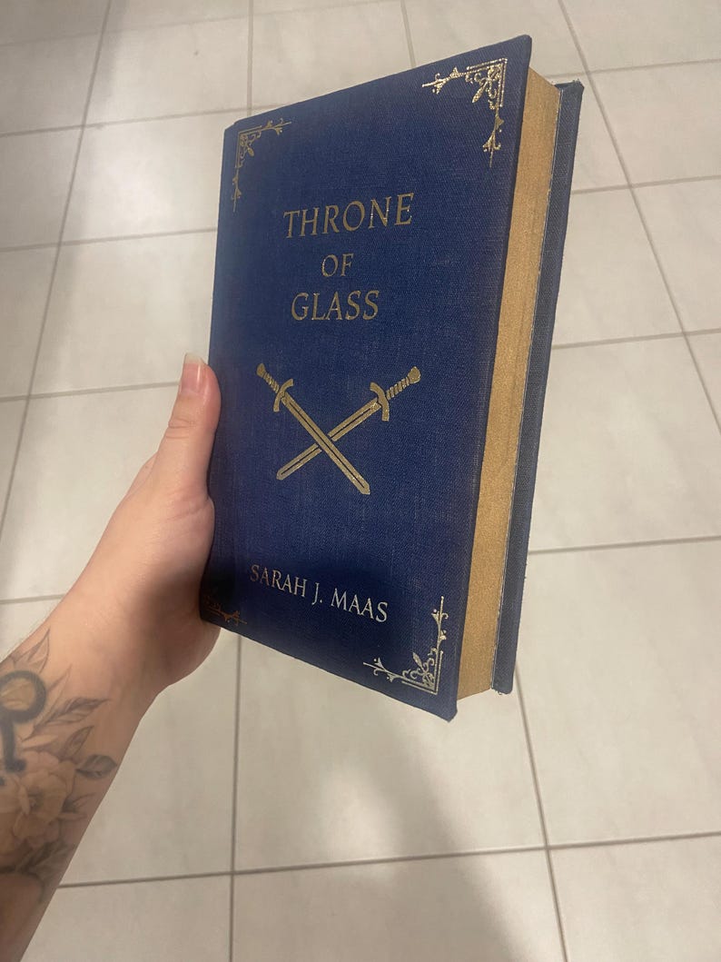 May include: A hardcover book titled "THRONE OF GLASS" by Sarah J. Maas. The book has a dark blue cover with gold lettering and decorative corner designs. Two crossed swords are featured below the title.