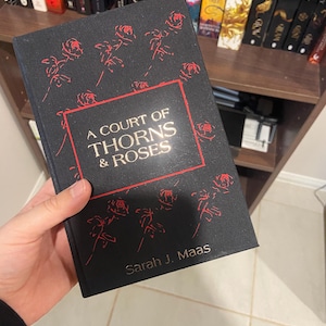 May include: A black book titled "A Court of Thorns & Roses" by Sarah J. Maas. The cover features red rose illustrations and text within a red border. The book is held in front of a bookshelf filled with other books.