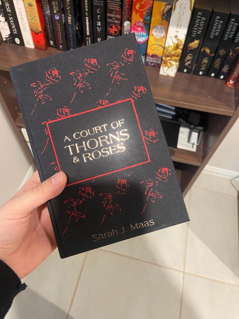 May include: A black book titled "A Court of Thorns & Roses" by Sarah J. Maas. The cover features red rose illustrations and text within a red border. The book is held in front of a bookshelf filled with other books.