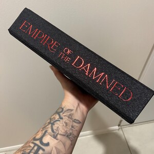 May include: A black book with the title "EMPIRE OF THE DAMNED" in red lettering. The book has a textured cover and is held at an angle, showcasing the title prominently.