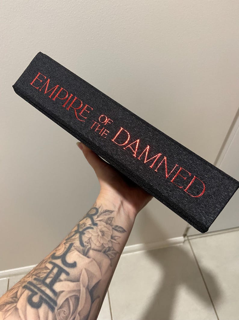 May include: A black book with the title "EMPIRE OF THE DAMNED" in red lettering. The book has a textured cover and is held at an angle, showcasing the title prominently.