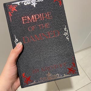 May include: A hardcover book titled "EMPIRE OF THE DAMNED" by Jay Kristoff. The book has a dark gray cover with red and silver decorative accents. The title is in red, and the author's name is at the bottom.