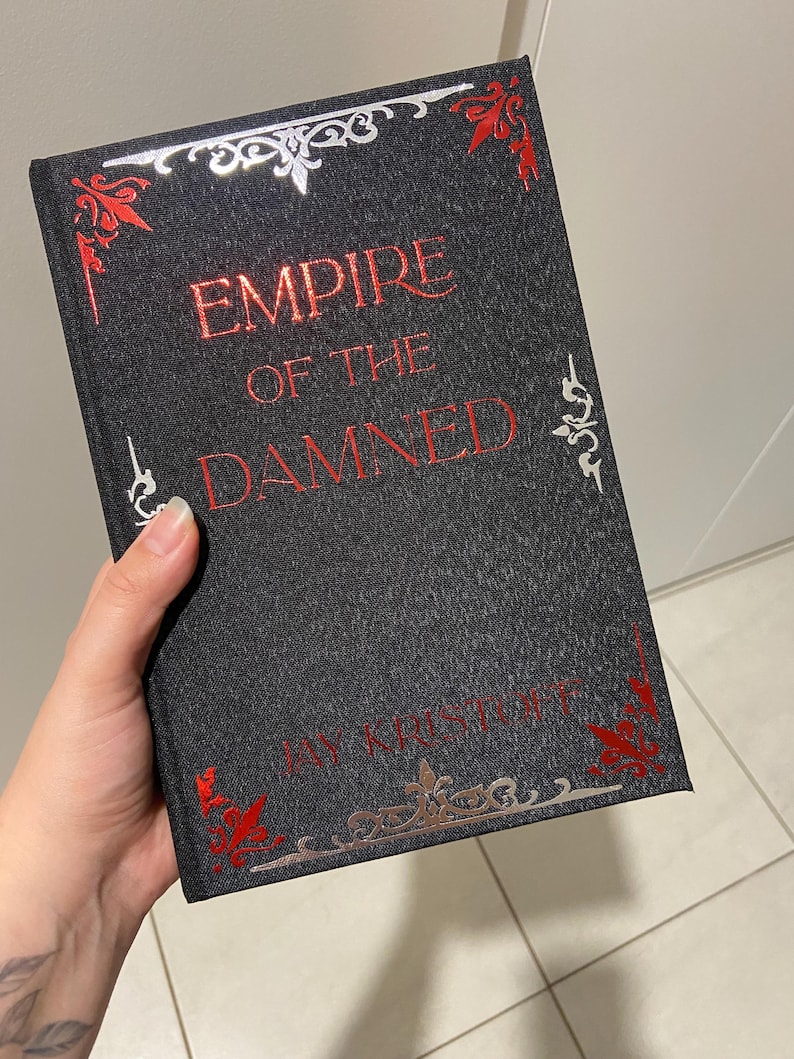 May include: A hardcover book titled "EMPIRE OF THE DAMNED" by Jay Kristoff. The book has a dark gray cover with red and silver decorative accents. The title is in red, and the author's name is at the bottom.