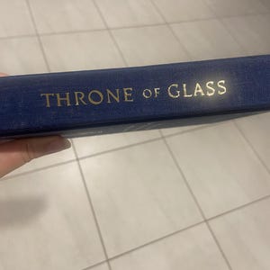 May include: A navy blue book with the title "THRONE OF GLASS" in gold lettering. The book's spine is visible, showcasing the title's embossed design. The book appears to be a fantasy novel.