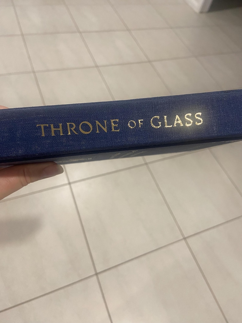 May include: A navy blue book with the title "THRONE OF GLASS" in gold lettering. The book's spine is visible, showcasing the title's embossed design. The book appears to be a fantasy novel.