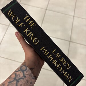 May include: A hardcover book titled "The Wolf King" by Lauren Palphreyman. The book has a black cover with gold lettering and a green accent on the spine. The book is held at an angle.