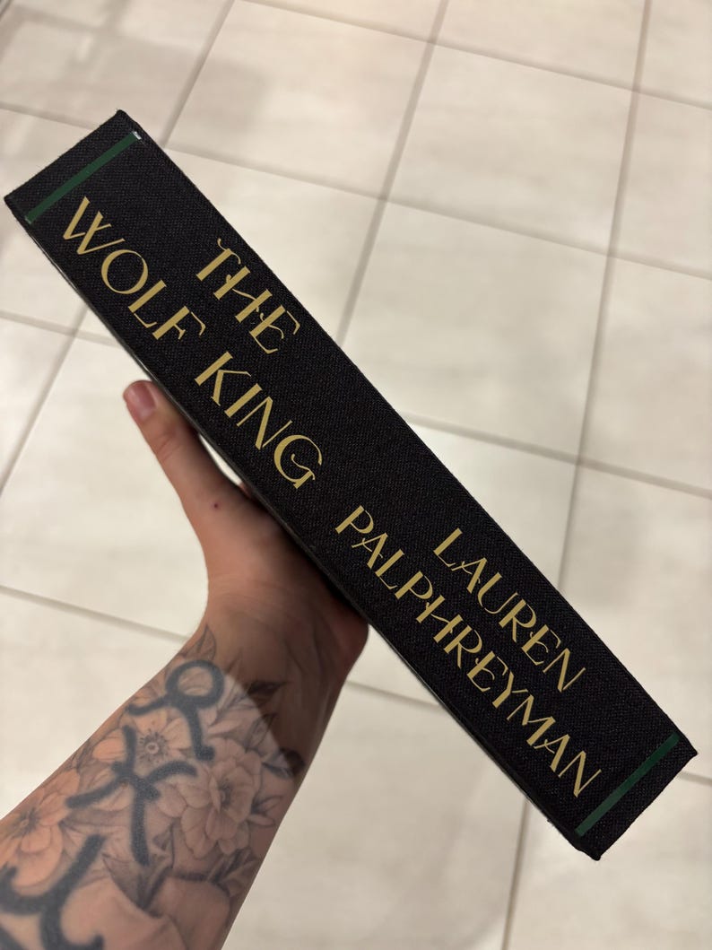 May include: A hardcover book titled "The Wolf King" by Lauren Palphreyman. The book has a black cover with gold lettering and a green accent on the spine. The book is held at an angle.