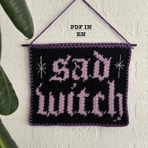 Könnte beinhalten: Ein schwarzer und lilafarbener gehäkelter Wandbehang mit dem Text "Sad Witch" in einer gotischen Schriftart. Der Behang hat einen lilafarbenen Rand und ist an einer Holzleiste befestigt.