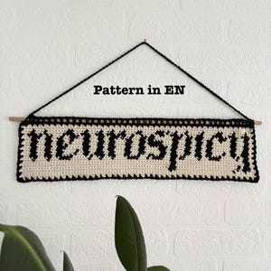 Pode incluir: Uma tapeçaria de parede de crochê creme com a palavra "neurospicy" em letras góticas pretas. A peça é emoldurada com uma borda preta e suspensa de um pino de madeira com uma corda preta. O texto "Pattern in EN" está acima.