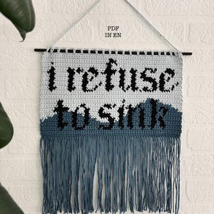 Könnte beinhalten: Ein gehäkelter Wandbehang mit dem Text "I refuse to sink" in schwarzer Wolle auf blau-weißem Hintergrund. Der Behang hat einen Fransenrand.
