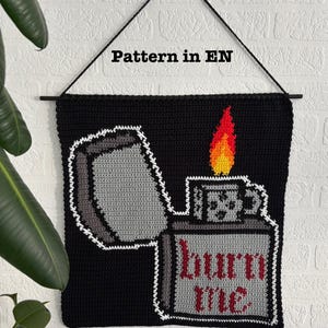 May include: Crocheted wall hanging featuring a black and white lighter with a red and yellow flame. The lighter has the text "burn me" on it.