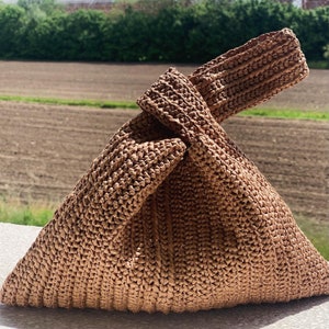 Könnte beinhalten: Eine braune gehäkelte Tragetasche mit einem geknoteten Henkel. Die Tasche ist aus einem gewebten Material gefertigt und hat eine dreieckige Form.