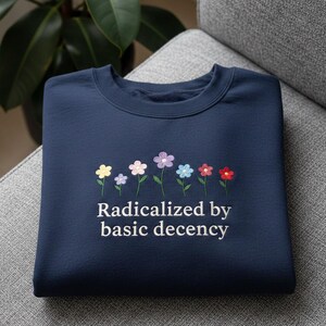 以下が含まれることがあります： 黄色、ピンク、紫、青、赤色の刺繍の花が施されたネイビーブルーのスウェットシャツ。「Radicalized by basic decency」の文字が花のすぐ下に刺繍されています。スウェットシャツは灰色の表面に折りたたまれています。